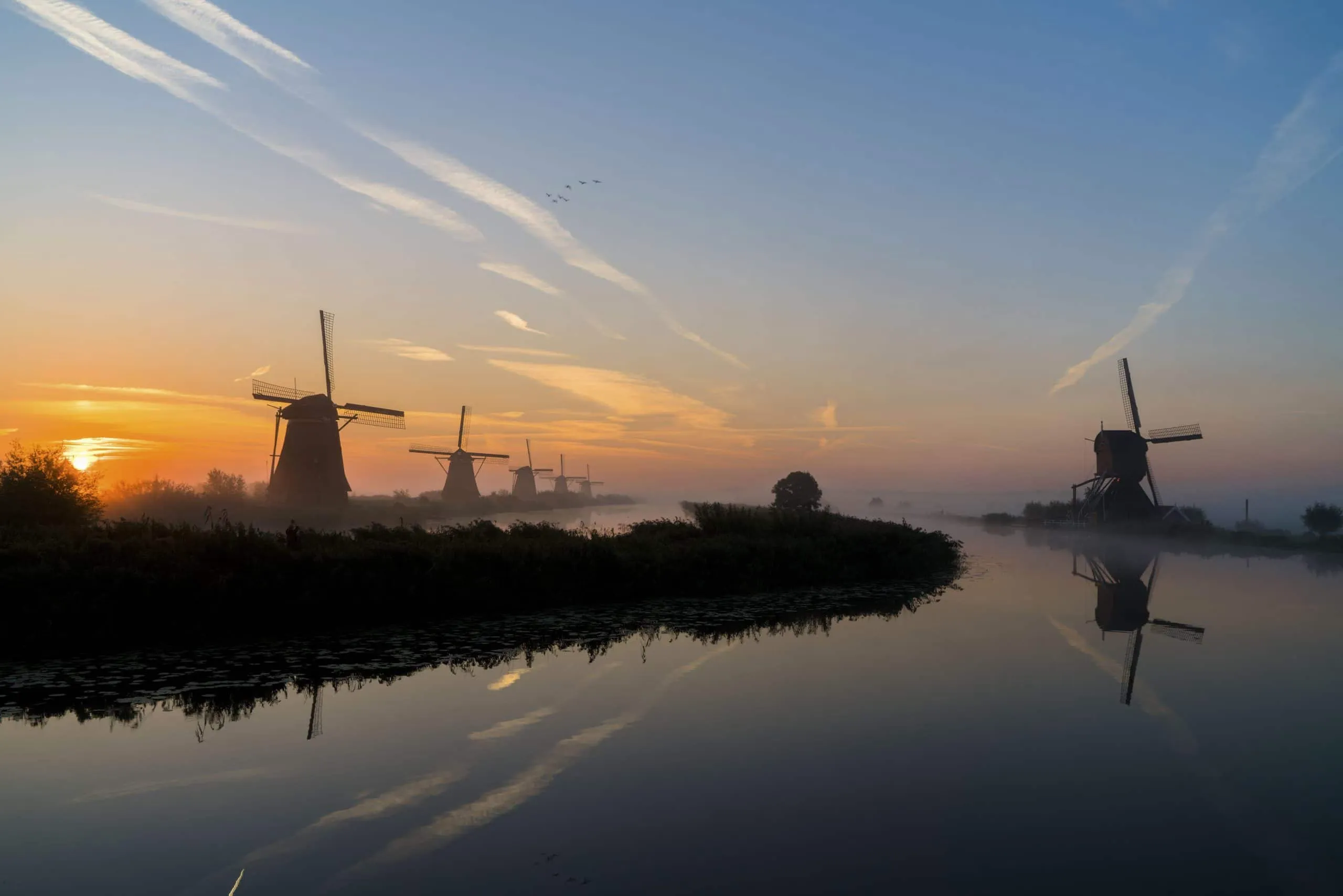 Silhouetted Dutch windmills at sunrise reflected in still polder water with mist over the fields
