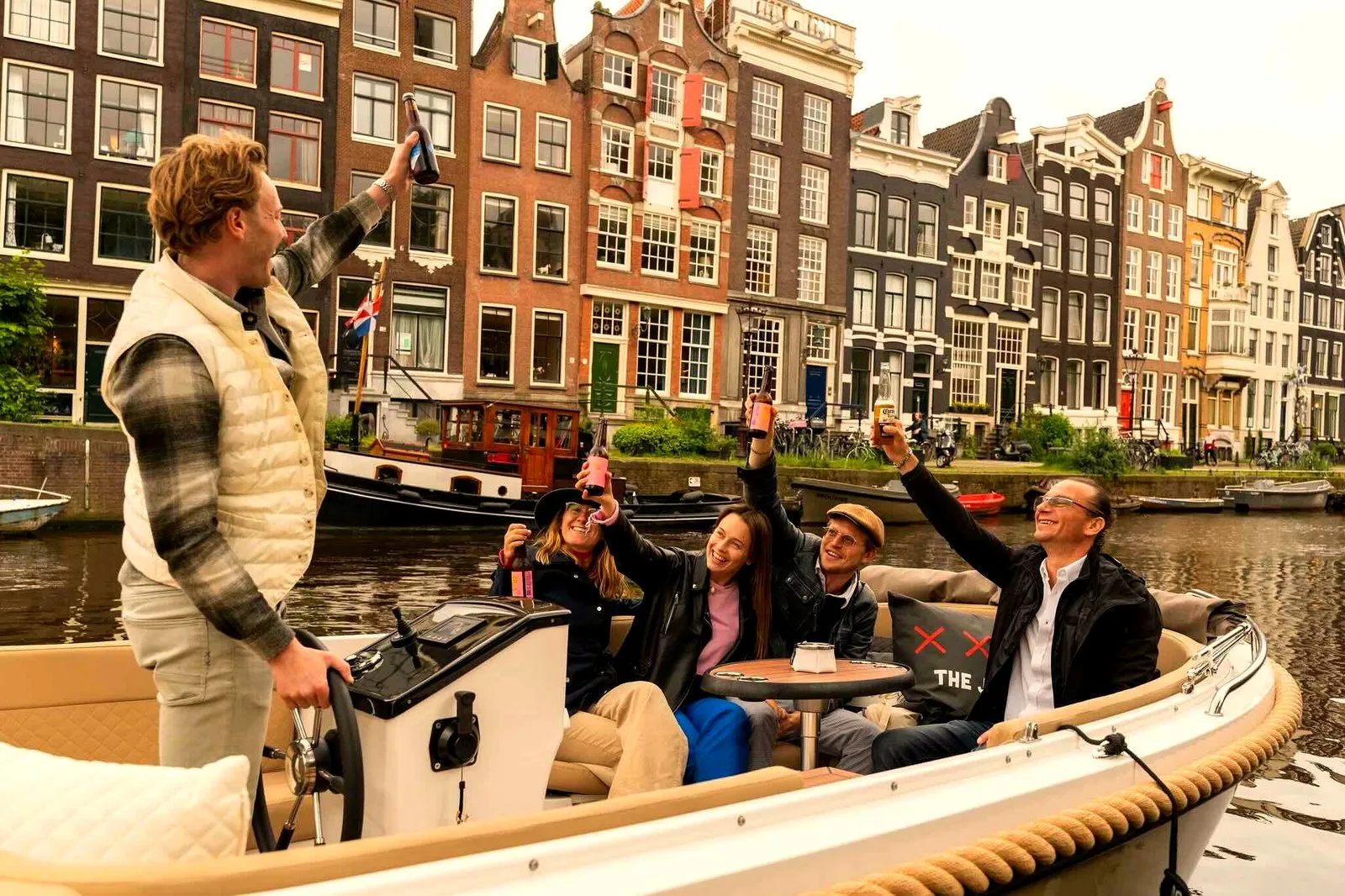 A host on an open canal boat pouring drinks while seated guests toast, classic Amsterdam canal houses behind