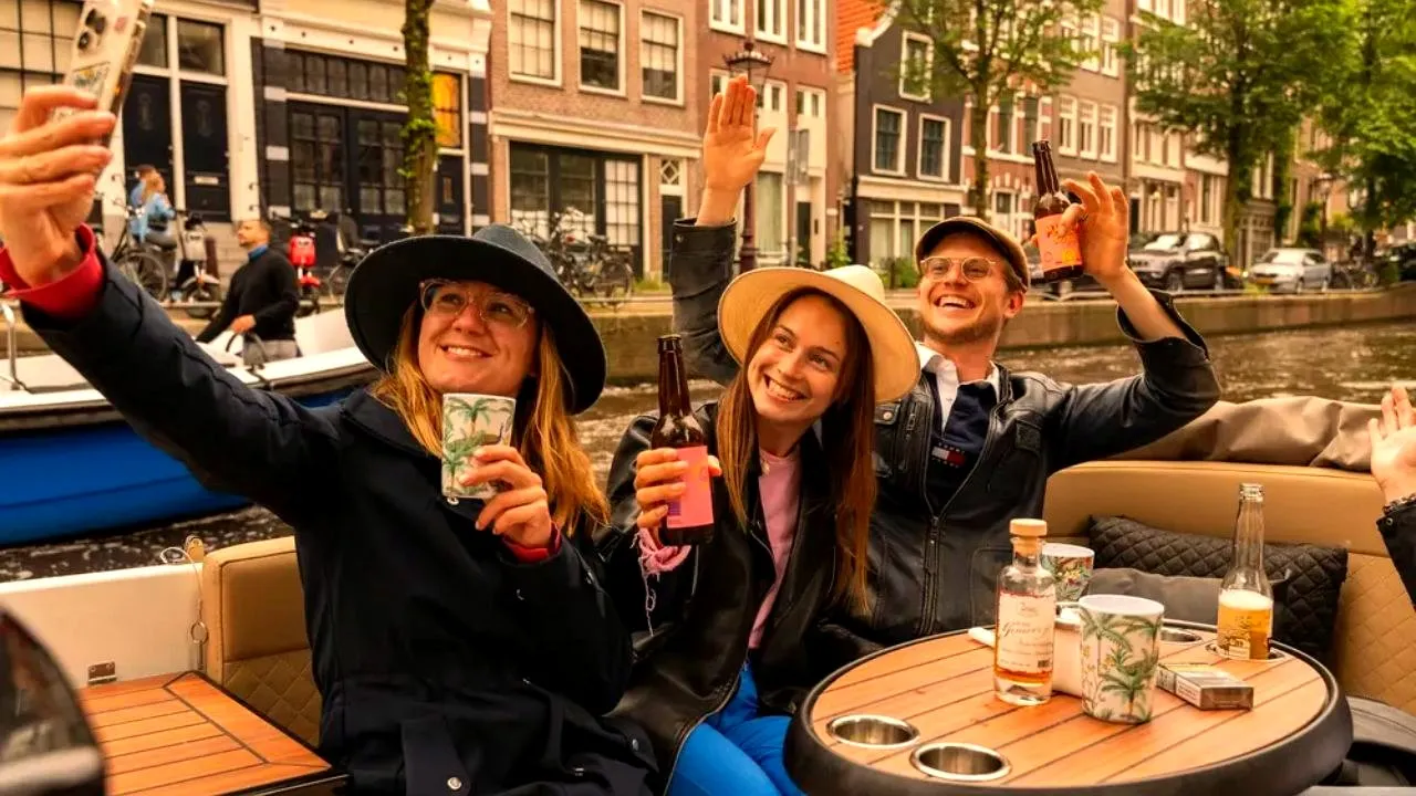 Three guests on an open boat in an Amsterdam canal taking a selfie while holding drinks