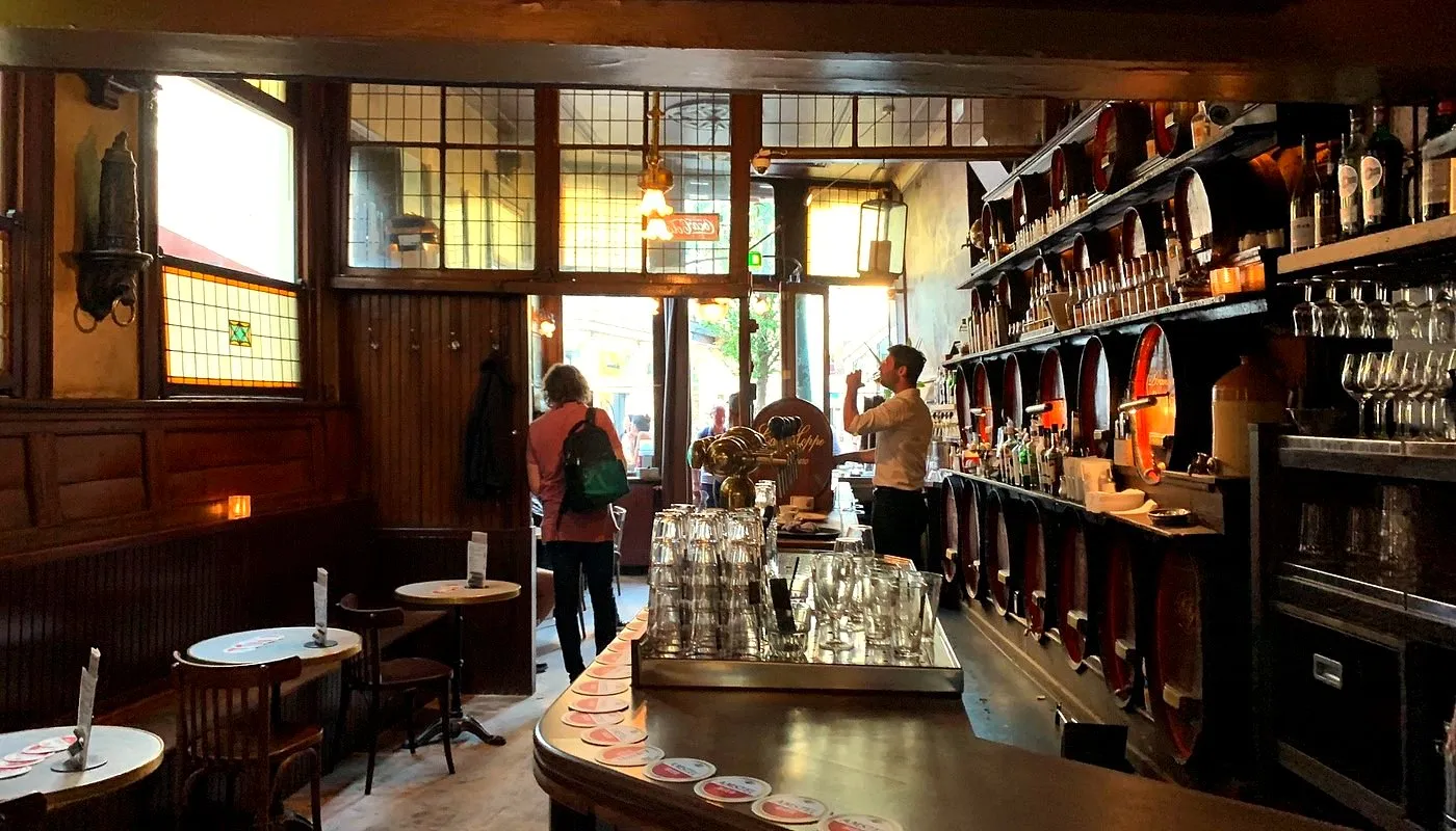 Dark wood-panelled historic Amsterdam pub with old liquor barrels on shelves, stained glass windows, marble tables and a bartender at work