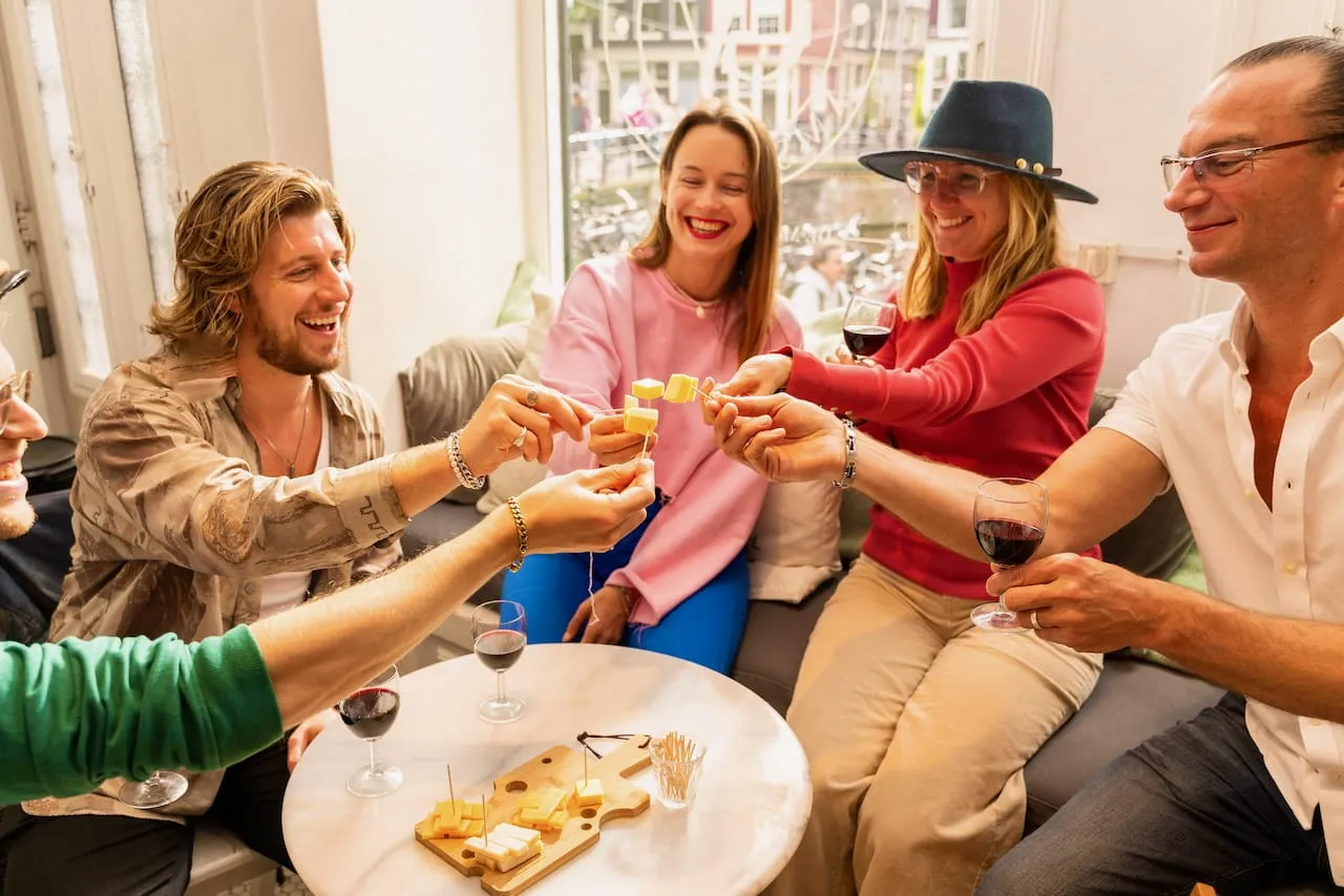 Four food tour guests laughing as they toast cubes of Dutch cheese on skewers over red wine by a canal window