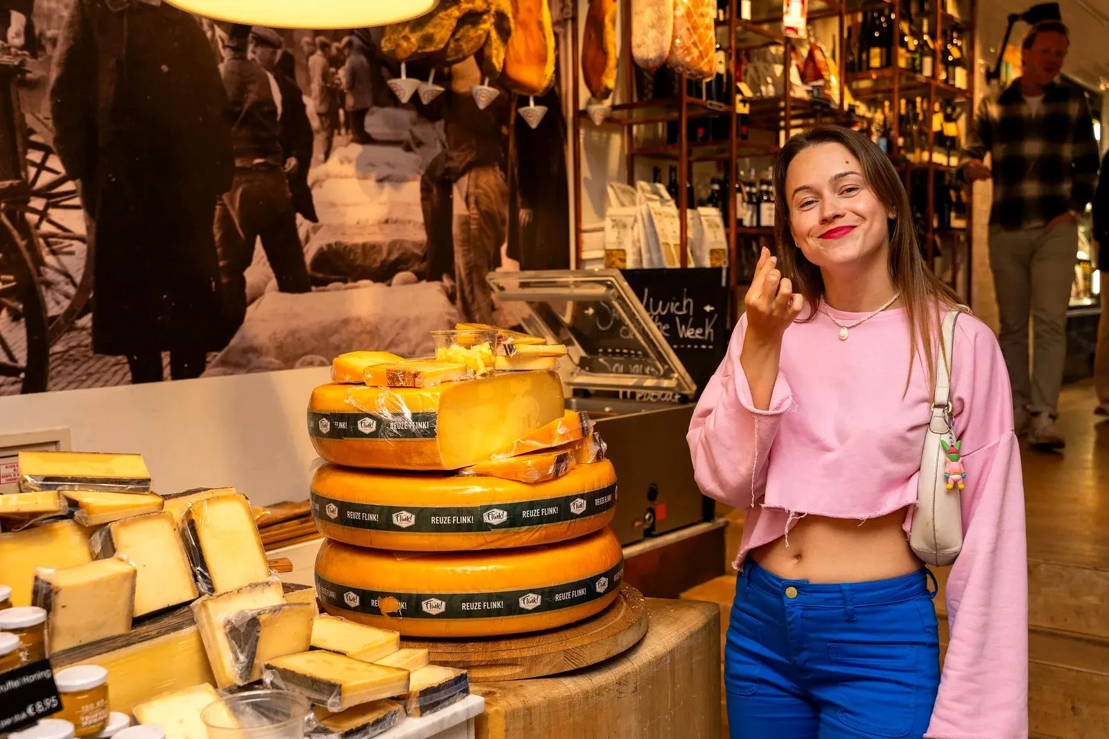 Woman in pink making a delicious gesture beside stacked cheese wheels in a shop