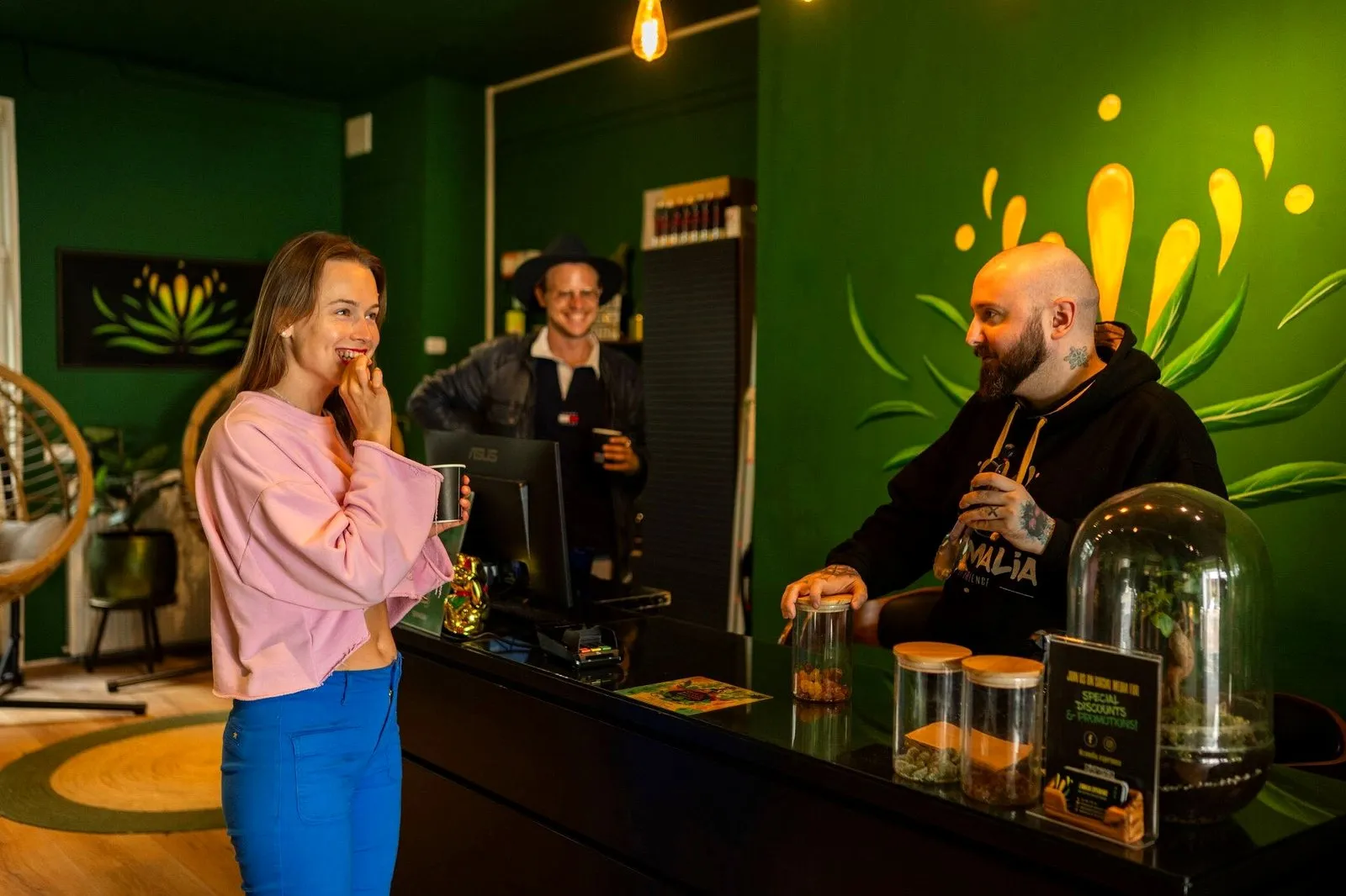 A guest smiling at the counter of a green Amsterdam herbal shop while the owner serves