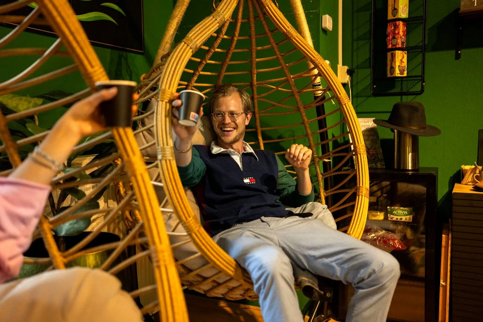 A guest toasting a cup from a hanging rattan chair in a cosy green Amsterdam shop
