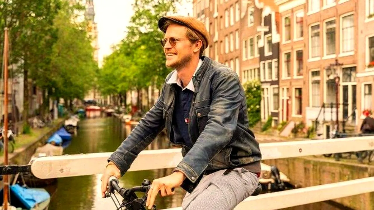 Smiling man in a flat cap riding a bike across a white wooden bridge over an Amsterdam canal