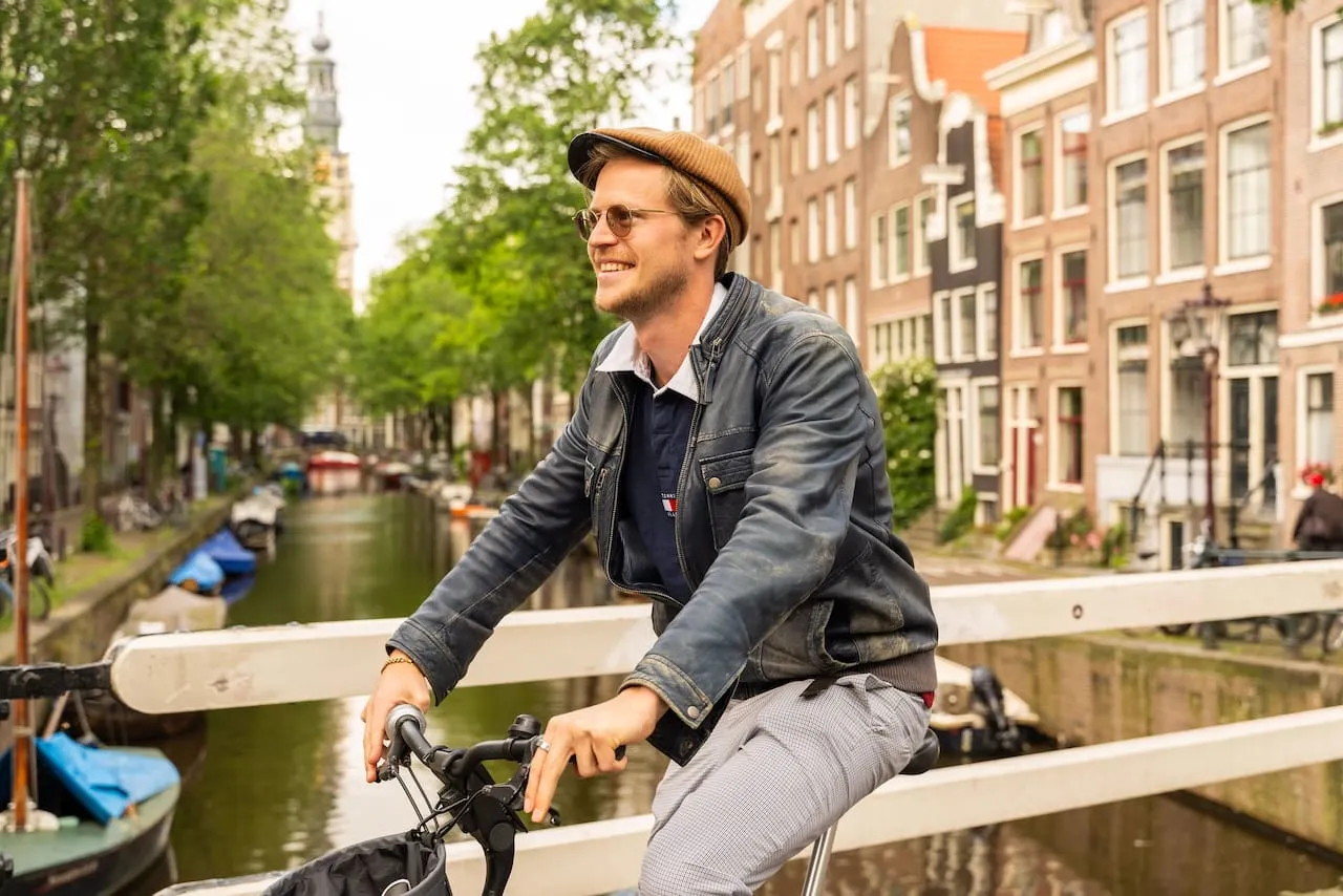 Smiling man in a flat cap riding a bike over an Amsterdam canal bridge with canal houses behind