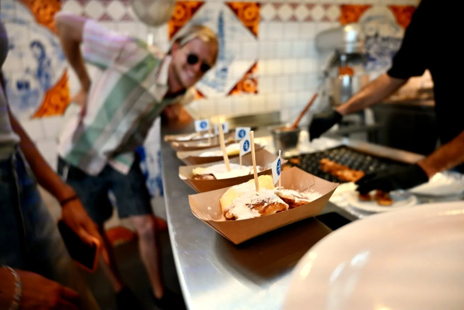 Trays of poffertjes with little flags being served at a tiled counter as a guest looks on