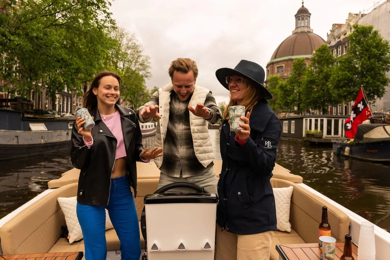 Three guests toasting drinks on a private Amsterdam canal boat with a domed church and canal houses behind