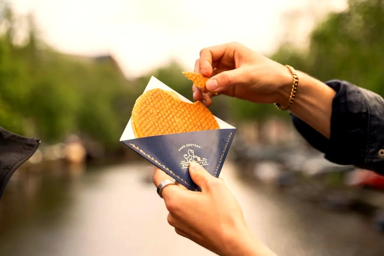 Hands sharing a stroopwafel in a branded sleeve above an Amsterdam canal