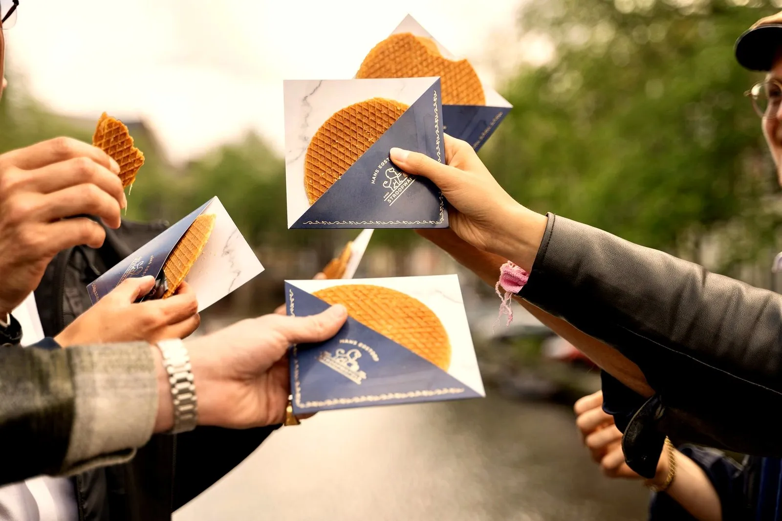 Several hands holding fresh stroopwafels in branded paper sleeves on an Amsterdam street