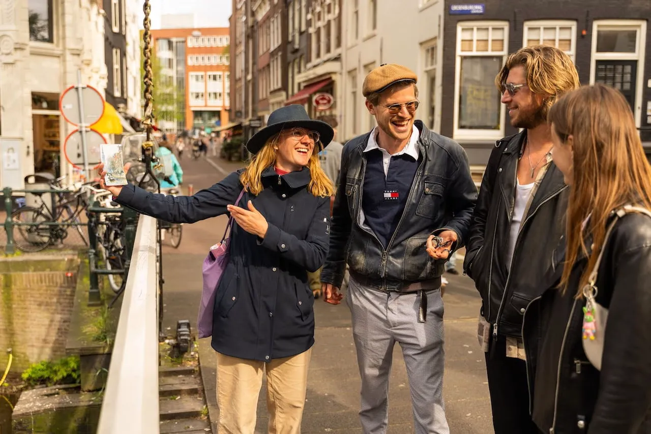 A tour guide in a hat and blue coat shows a small card to three guests on an Amsterdam street bridge