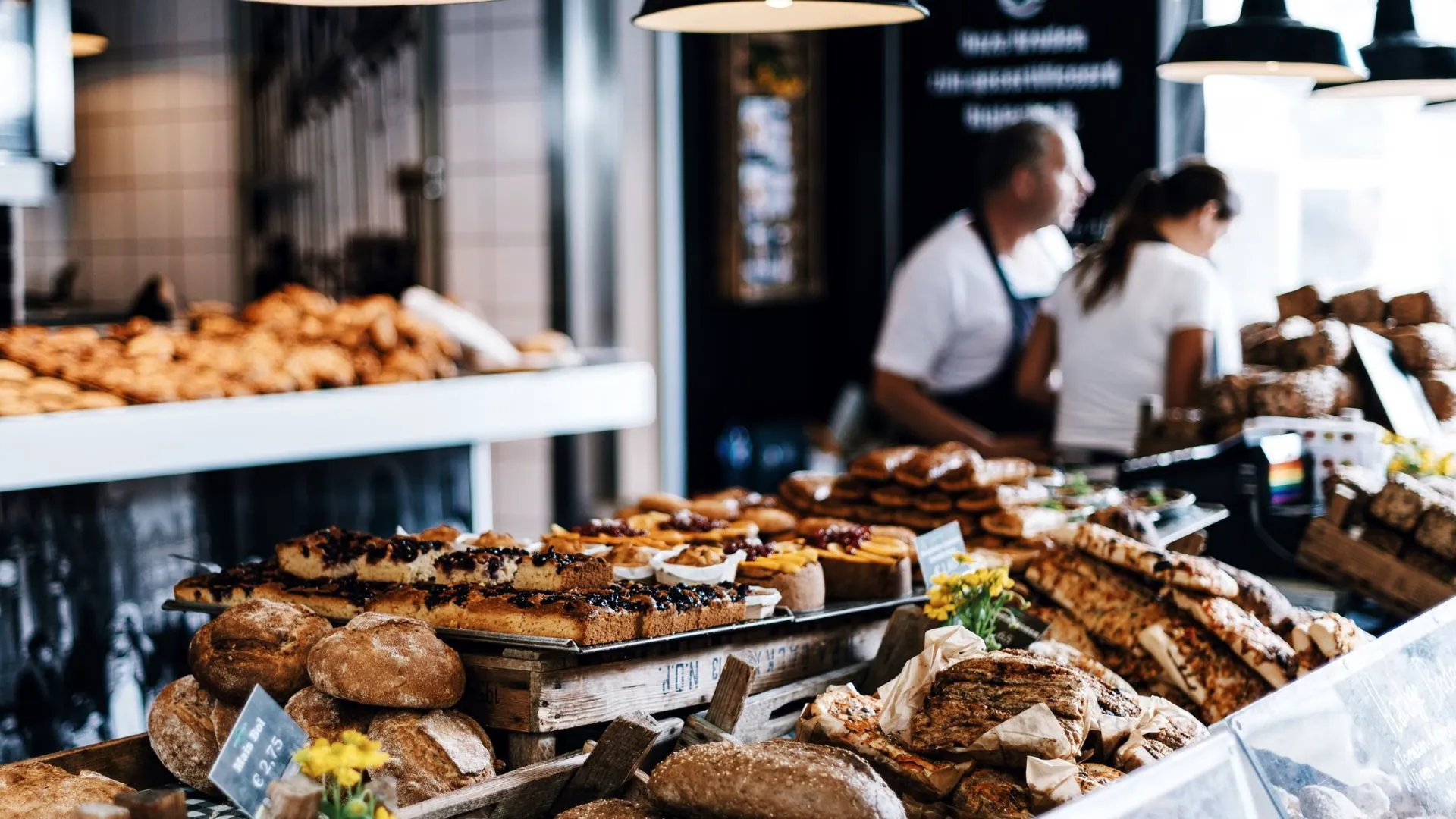 Bakery counter piled with rustic loaves, seeded breads and fruit pastries, two staff members working behind