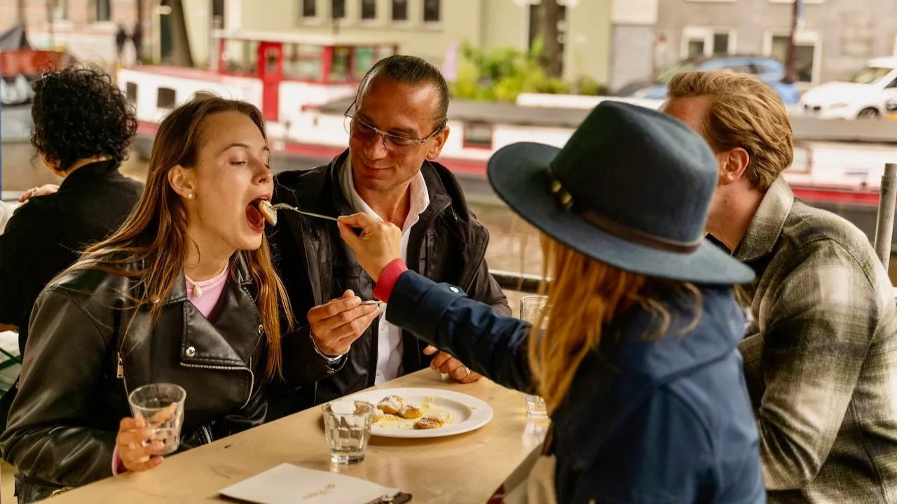 Friends sharing dessert at an outdoor canalside table in Amsterdam, one being fed a forkful
