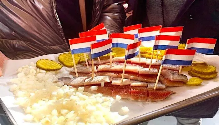 Tray of raw herring pieces on cocktail sticks with small Dutch flags, served with chopped onions and pickles
