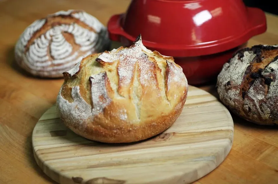 Three rustic round loaves of crusty bread on a wooden counter, one on a cutting board in front of a red ceramic baking pot