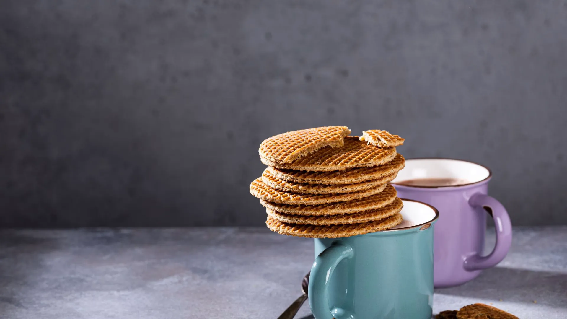 Stack of stroopwafels balanced on a turquoise enamel mug with a purple mug of coffee behind, against a gray backdrop