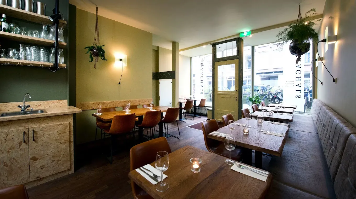 Bright restaurant interior with olive green walls, wooden tables set with wine glasses and hanging plants near the entrance