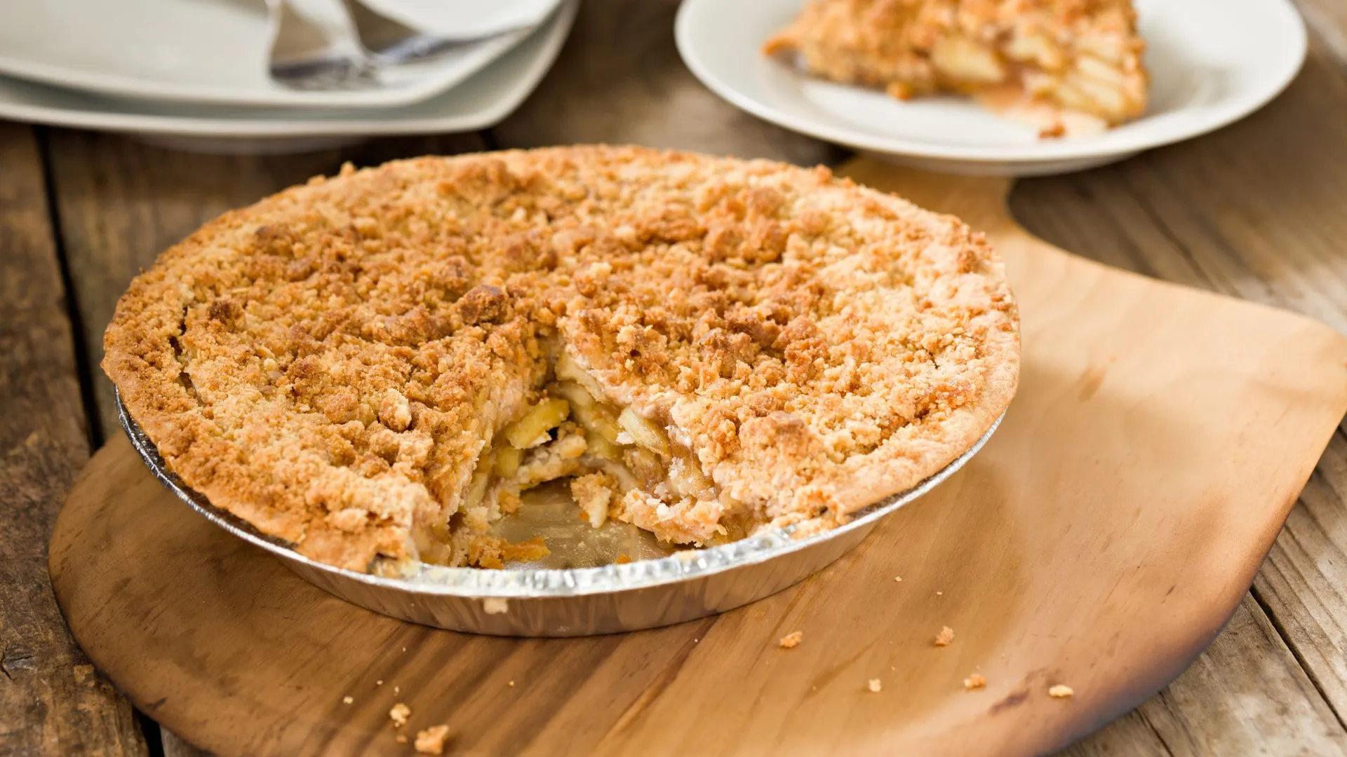 Whole apple pie with a golden crumble topping in a foil tin, one slice removed, on a wooden board