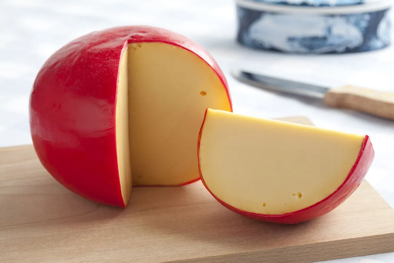 Round Edam cheese in red wax with a wedge cut out, on a wooden board with a cheese knife behind