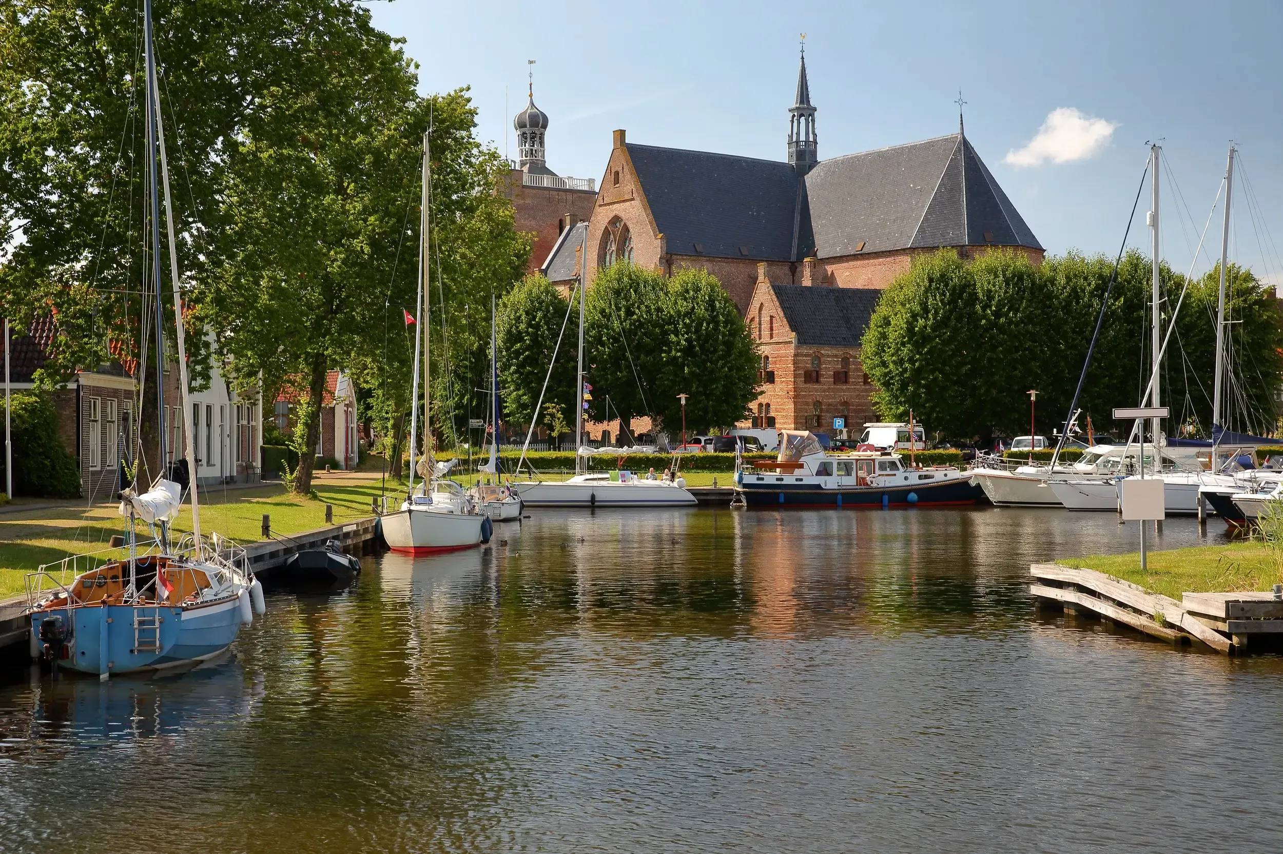 Sailboats moored in a quiet Dutch village harbor with a large brick church and green trees behind