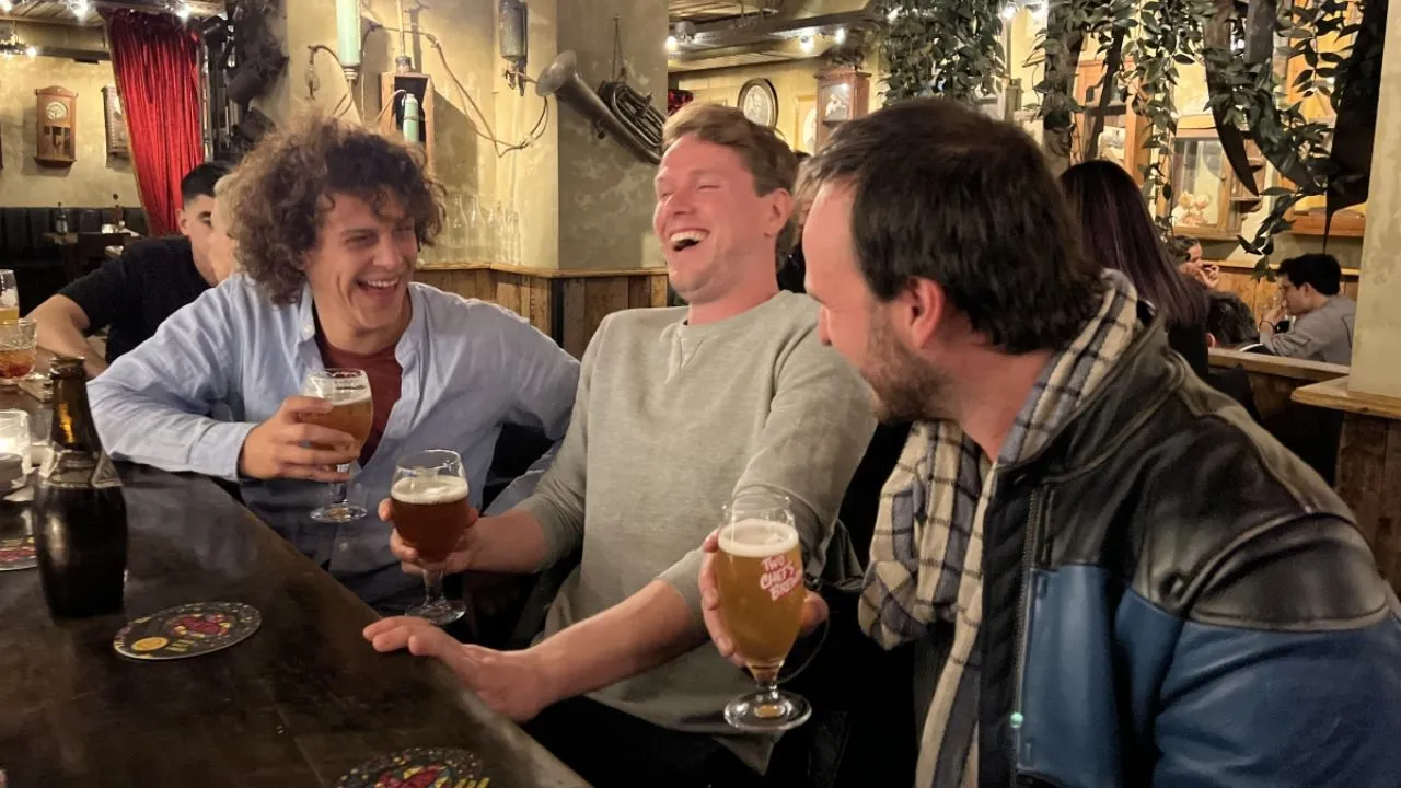 Three men laughing together over glasses of beer at the wooden bar of a cozy brown cafe