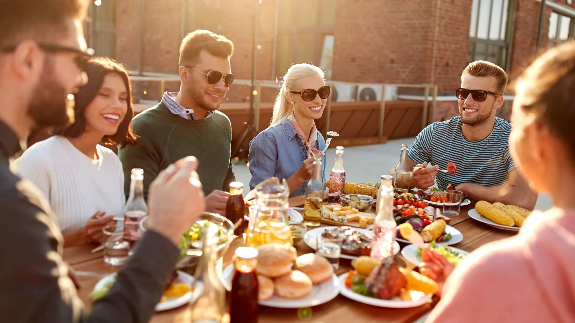 Group of friends in sunglasses sharing plates of food and drinks around a wooden table on a sunny rooftop