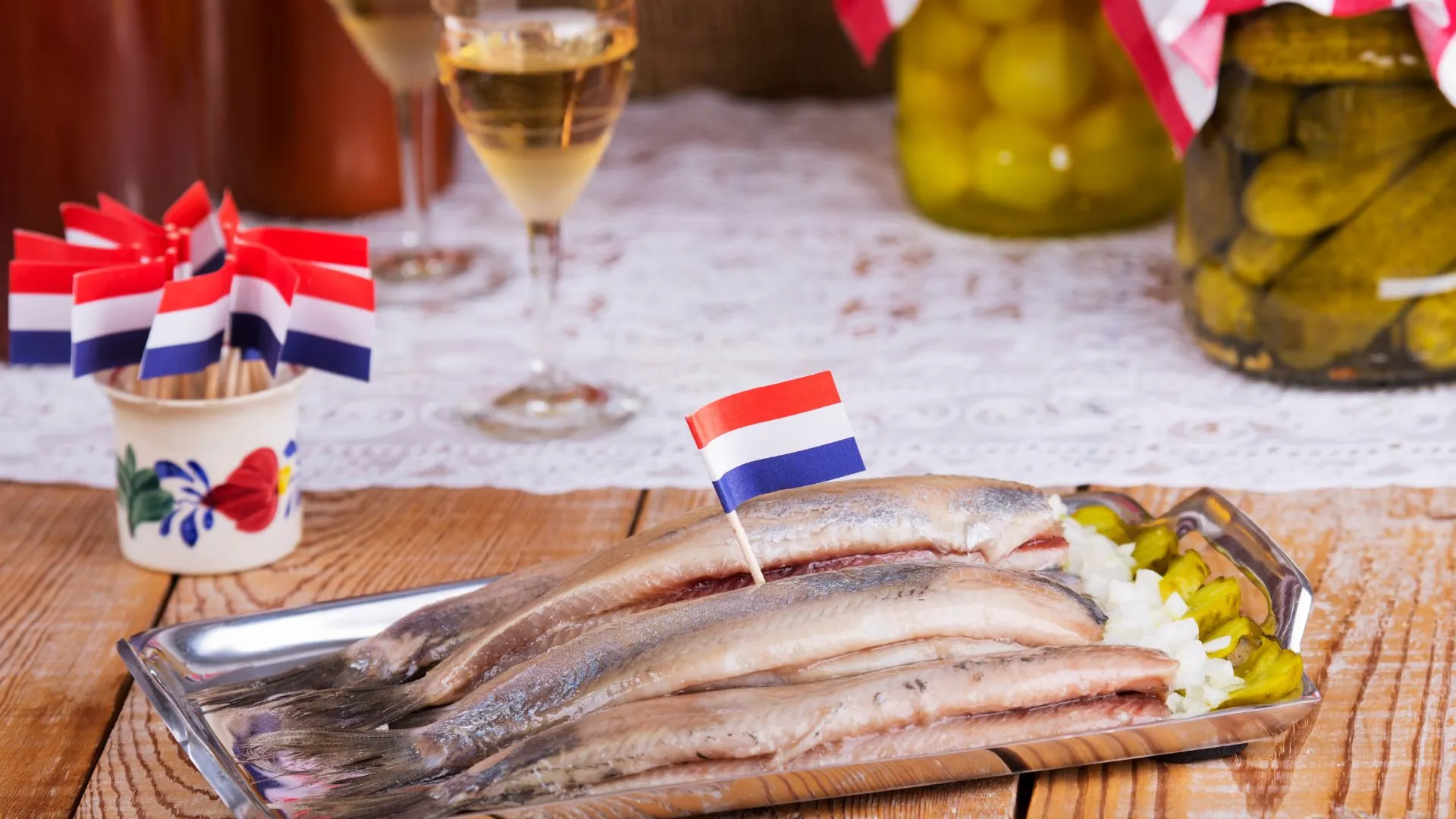 Raw herring fillets on a metal tray with chopped onions, pickles, and small Dutch flags on toothpicks
