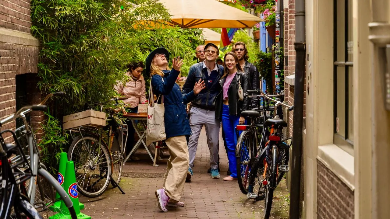 Tour group looking up as their guide points out details in a narrow, plant-filled Amsterdam alley lined with bikes