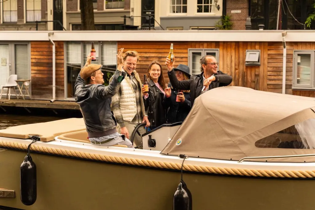 Five friends on an open sloop raise beer bottles in a toast, with wooden houseboats along the Amsterdam canal behind them