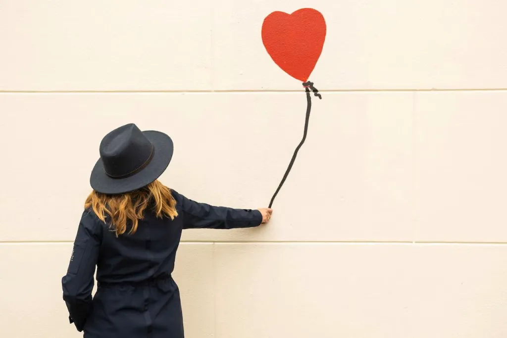 Woman in a dark hat seen from behind reaches toward a red heart balloon painted on a cream wall, as if holding its string