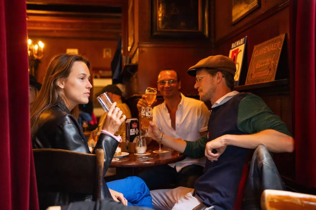 Three people drink beer around a small wooden table in a dark wood-paneled Amsterdam brown cafe with old paintings and enamel signs