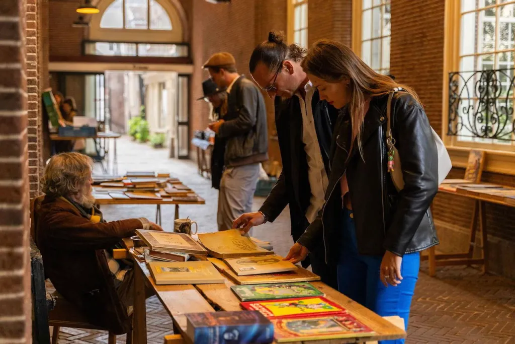 Couple browses old books at a wooden stall in a brick arcade book market in Amsterdam while the seller looks on
