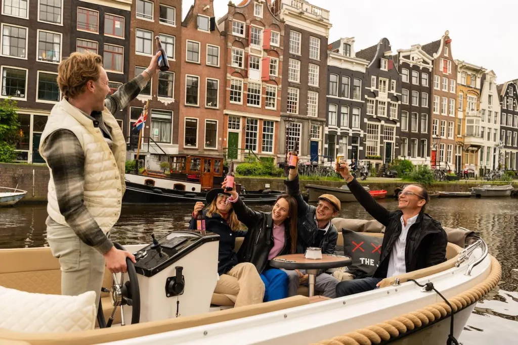Skipper and four guests raise drinks in a toast aboard an open boat, with tall Amsterdam canal houses lining the water behind