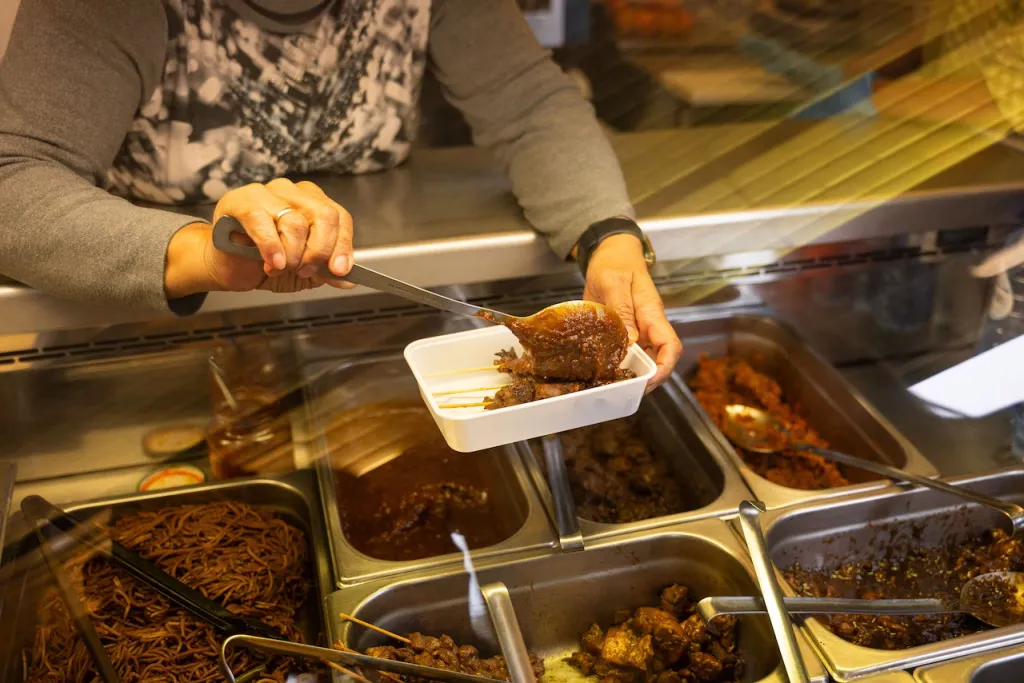 Server spoons a portion of stewed meat into a takeaway container over steel trays of noodles and stews at a Surinamese counter