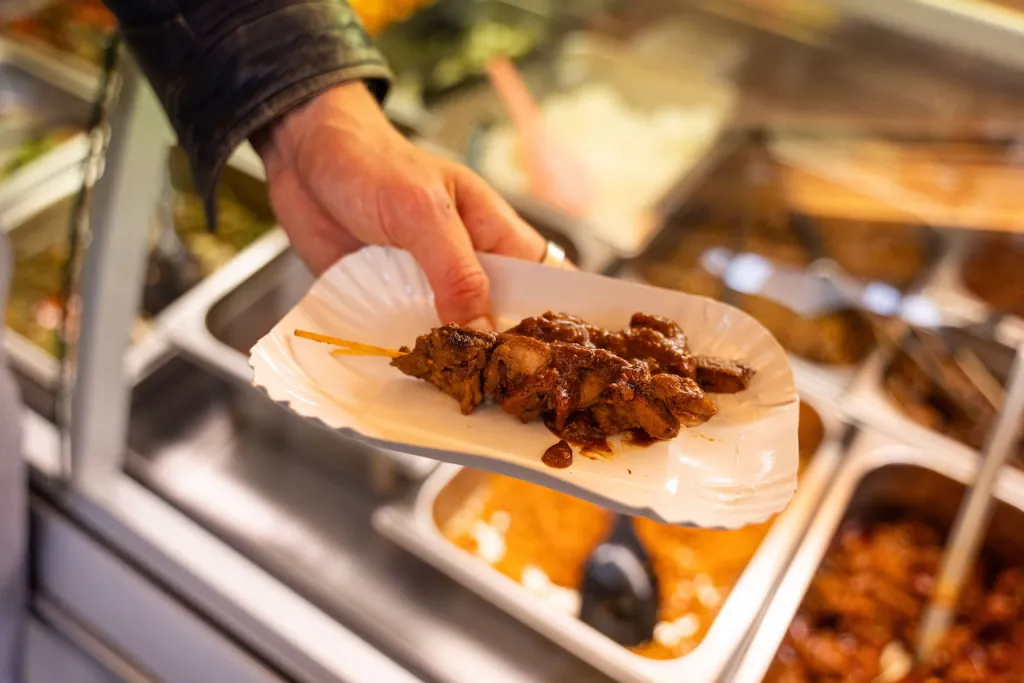 A hand holds a paper tray with a saucy chicken satay skewer over a food counter filled with warm dishes