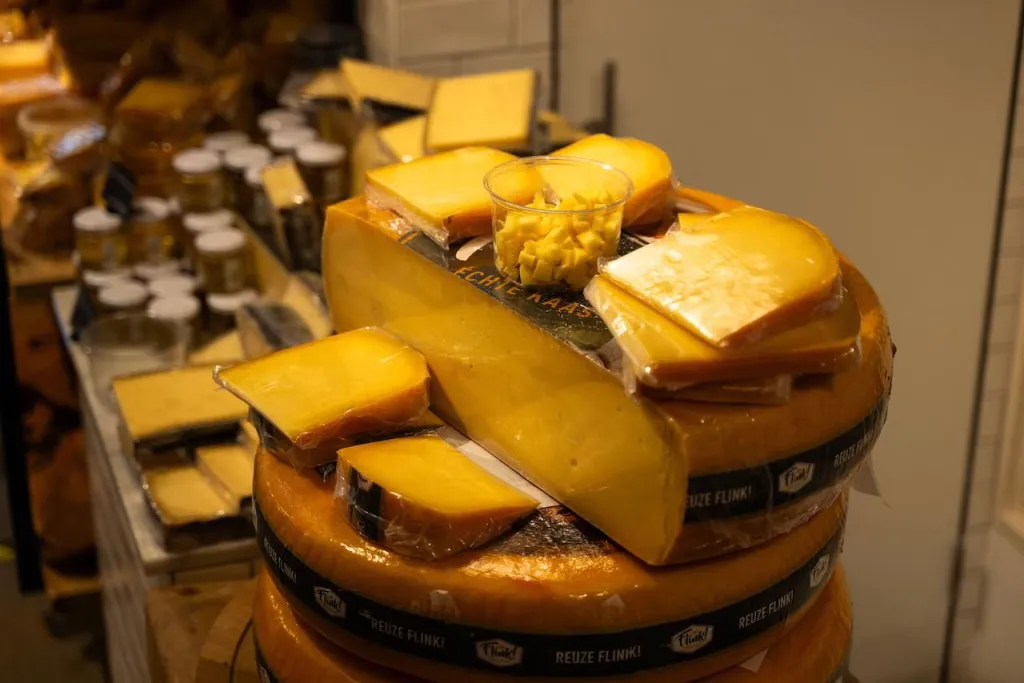 Stacked Gouda cheese wheels and wedges in a shop with a cup of sample cubes on top