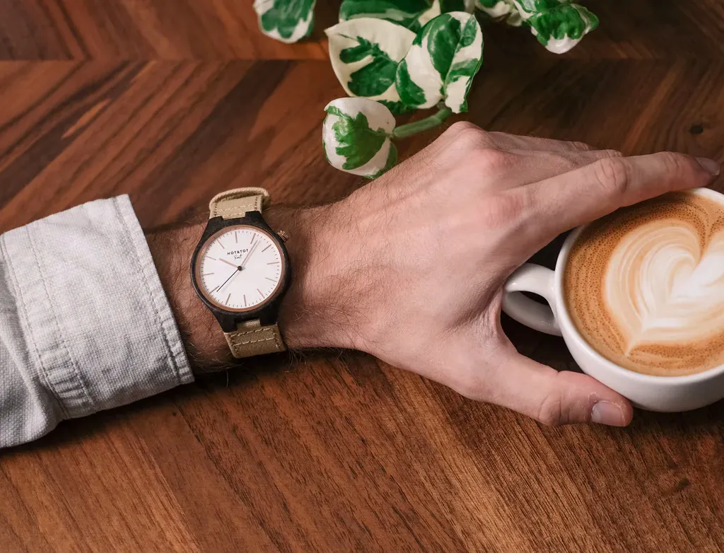 A man's wrist with a watch reaches for a latte with heart-shaped foam art on a wooden table beside a houseplant