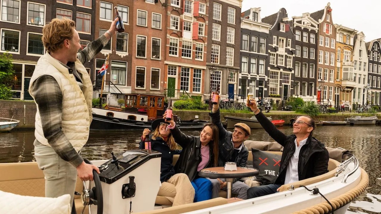 Skipper raising a bottle while four guests toast with drinks on a private boat in front of Amsterdam canal houses