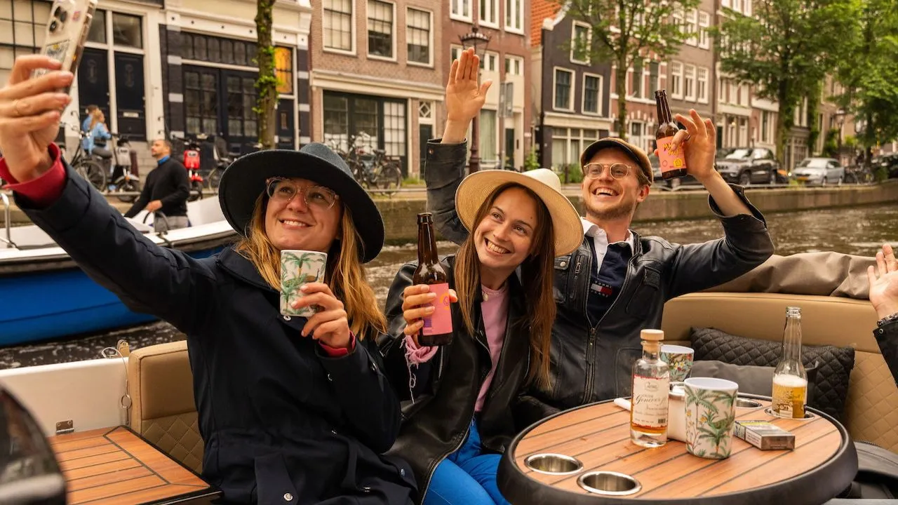 Three guests in hats taking a selfie and waving with bottles and cups on a boat cruising an Amsterdam canal