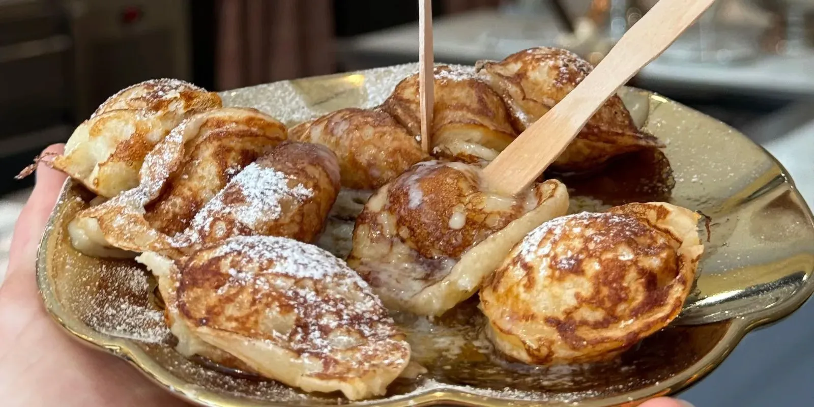 Hand holding a gold plate of fresh poffertjes dusted with powdered sugar and melted butter, with wooden forks stuck in