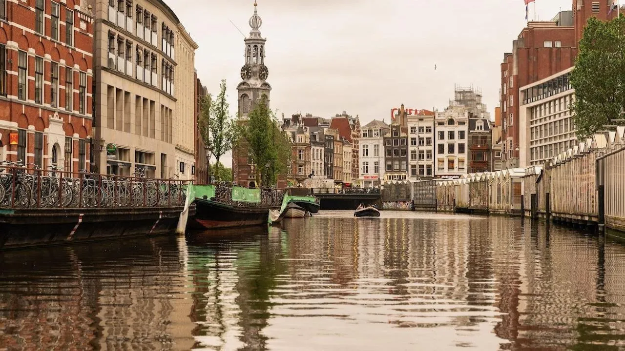 View from the water of an Amsterdam canal leading toward the Munttoren clock tower with moored barges along the quay