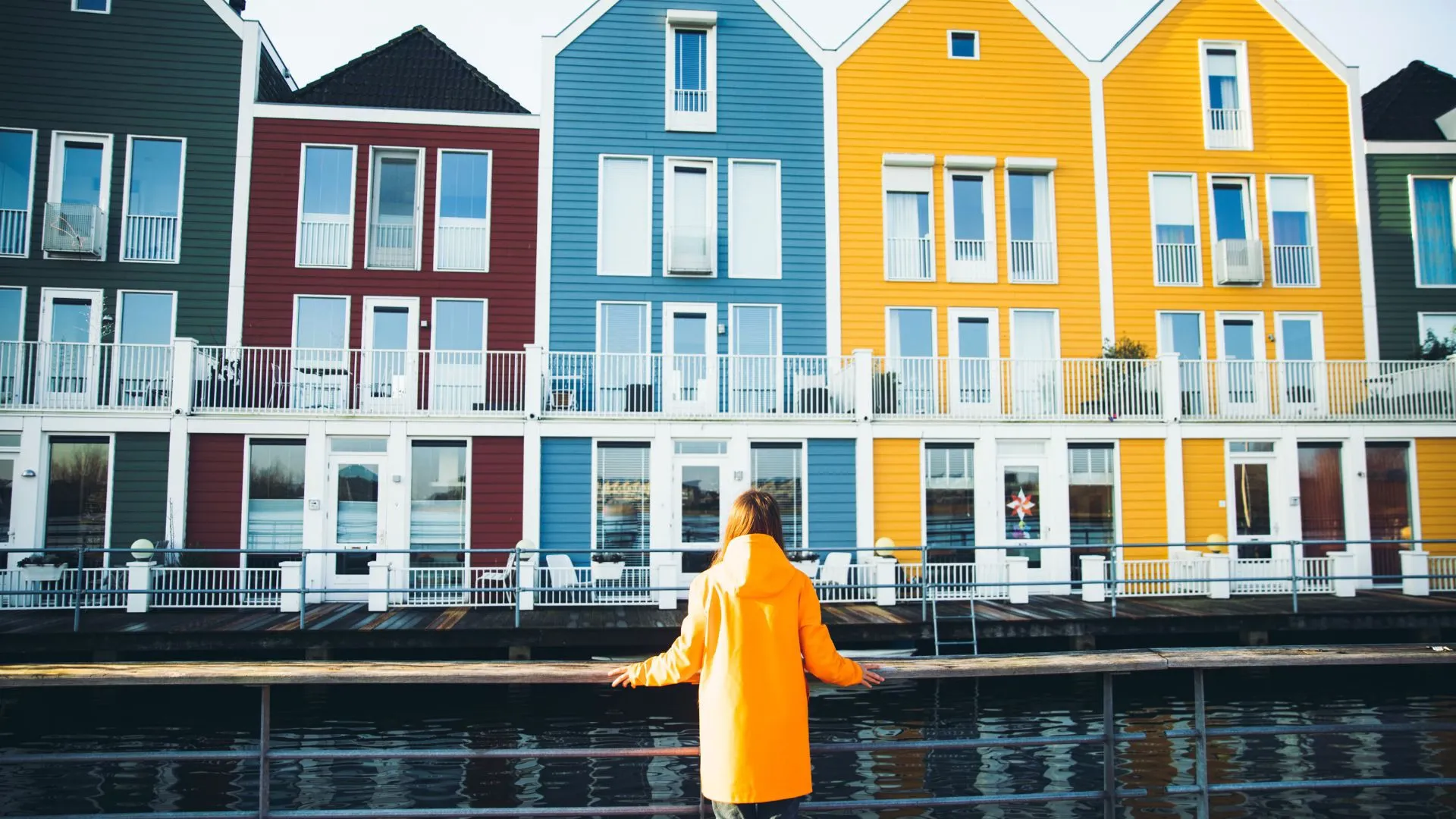 Woman in a yellow raincoat standing at a waterside railing in front of a row of colorful wooden Dutch houses
