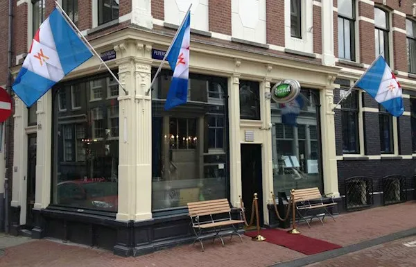 Corner restaurant in a brick Amsterdam building with blue and white flags above the entrance, wooden benches and a red carpet at the door