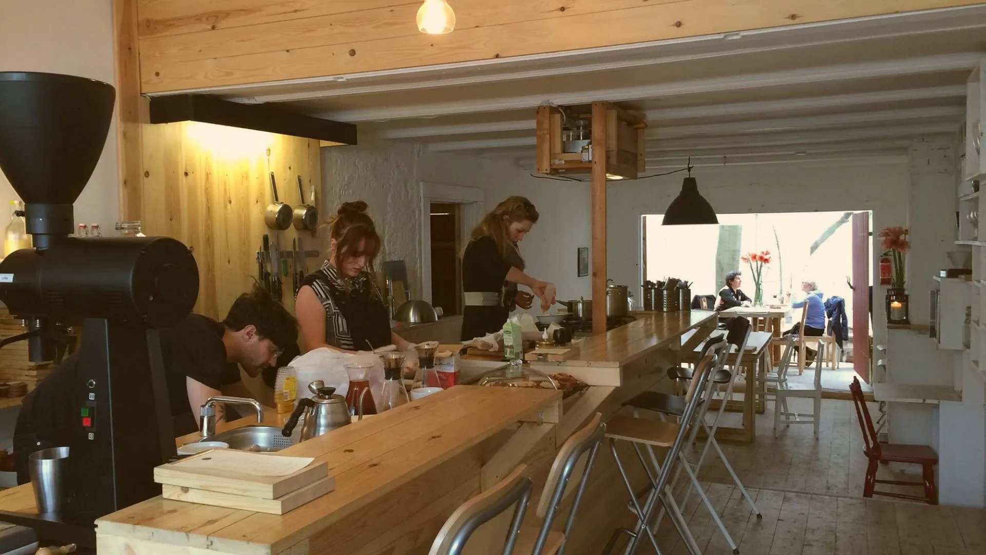 Light wood cafe interior with baristas working behind a long counter, a black coffee grinder in the foreground and guests seated near the window