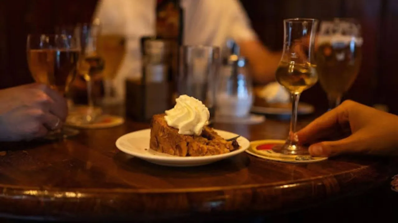 Slice of Dutch apple pie with whipped cream on a wooden pub table, next to a tulip glass of jenever and glasses of beer