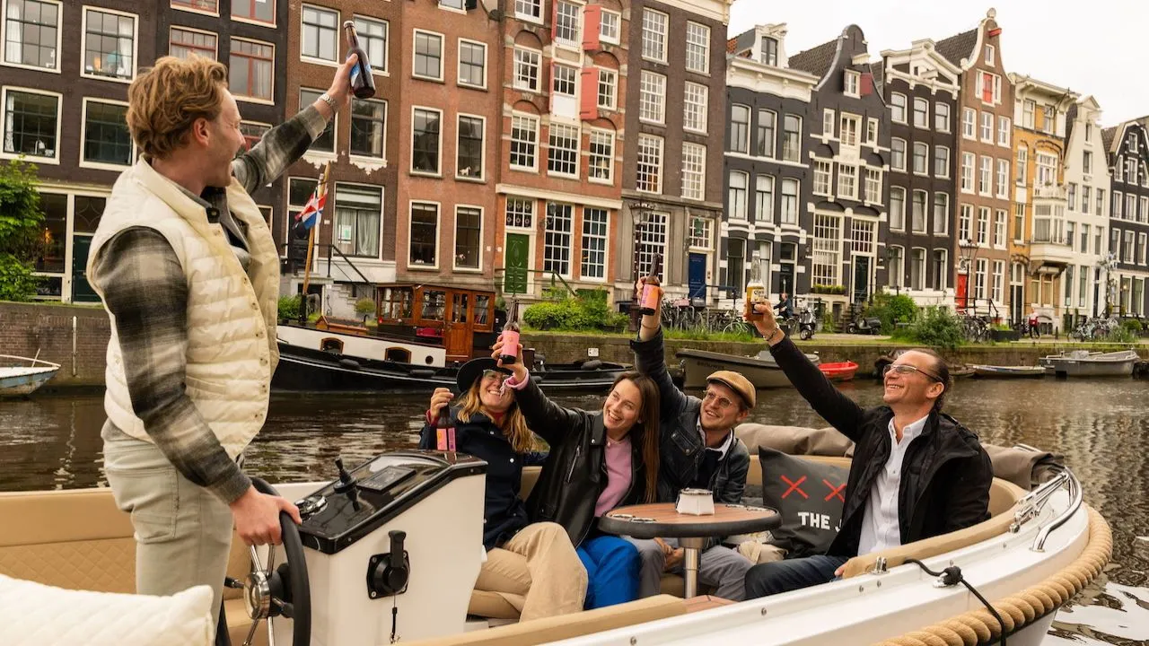 Group on an open boat raising drinks in a toast while the skipper holds up a bottle, with Amsterdam canal houses behind them