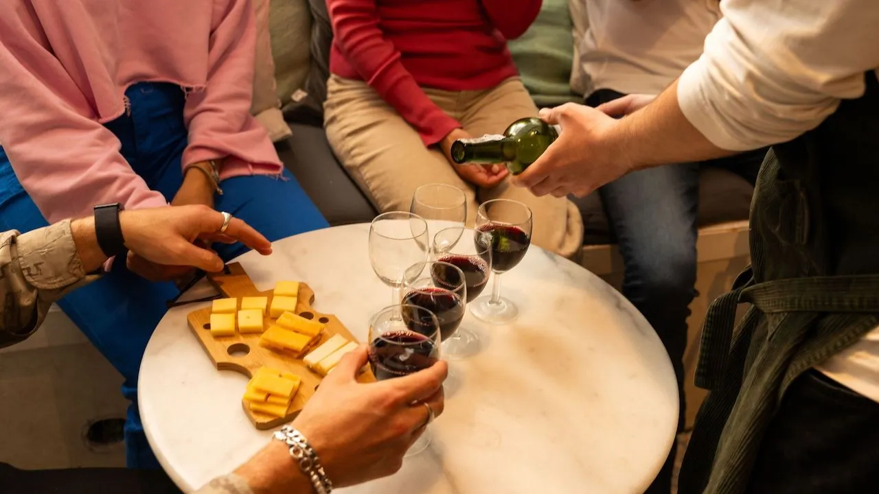 Host pours red wine into glasses on a small marble table set with a wooden board of Dutch cheese cubes while guests reach for a taste