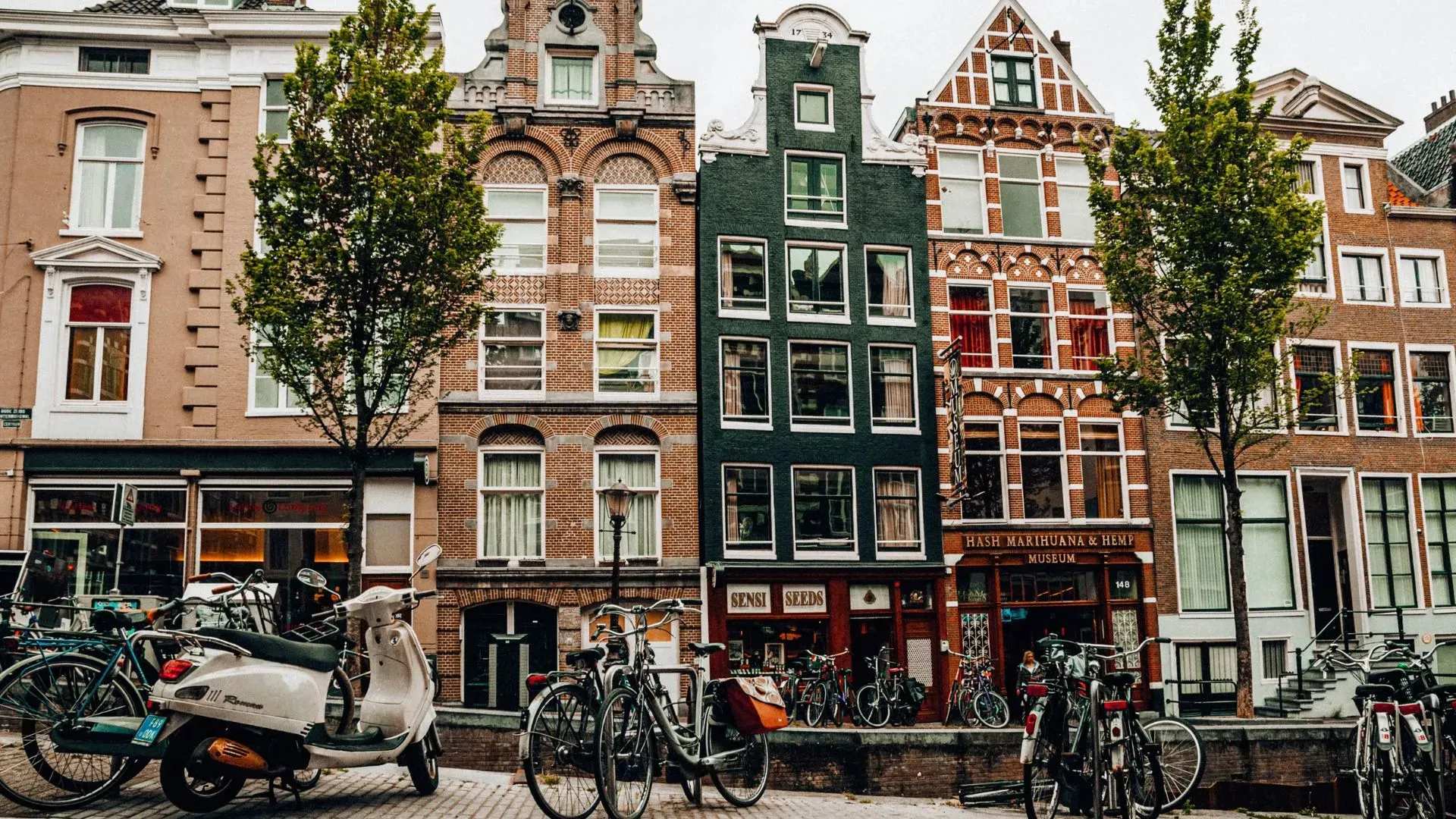 Row of narrow Amsterdam canal houses in brick and dark green, with bicycles and scooters parked along the street