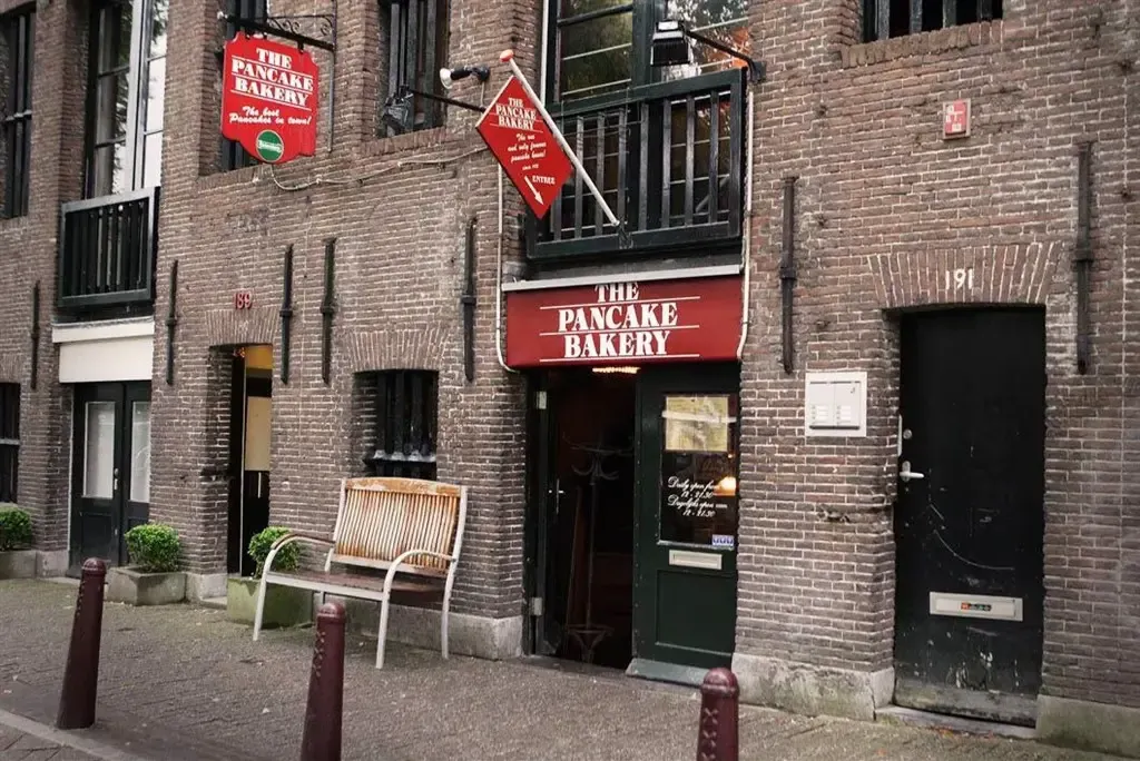 Brick canal-house facade with red signs for The Pancake Bakery, a white bench beside the dark green entrance