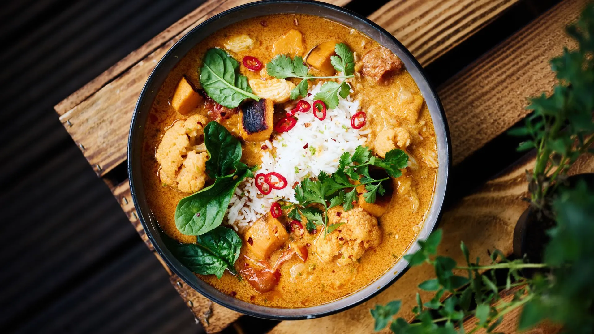Overhead view of a bowl of creamy vegetable curry with cauliflower, rice, fresh herbs and red chili slices on a wooden crate